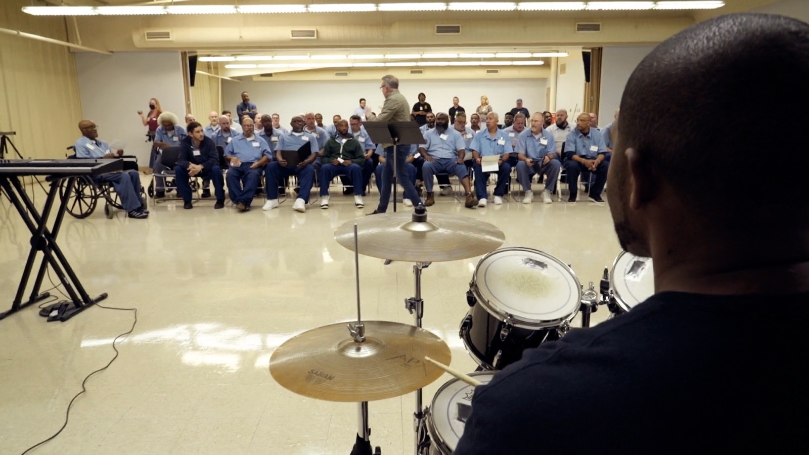 Seen over a drummer's shoulder, the seated choir faces their conductor during rehearsal.