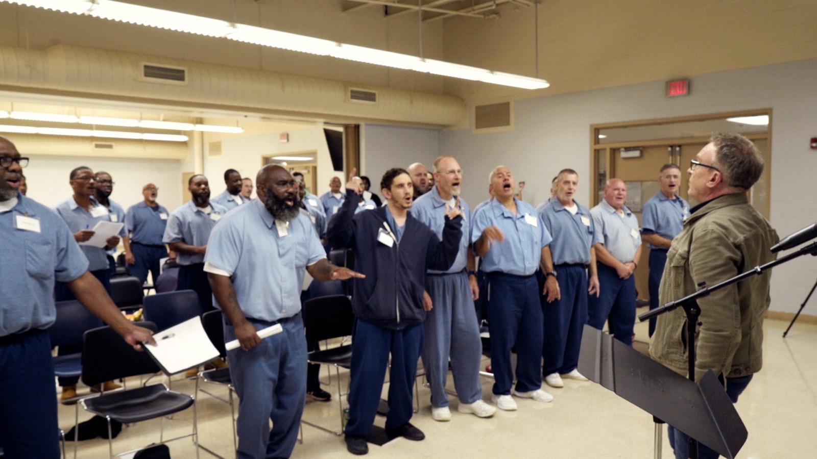 The choir mid-song in a bright rehearsal room, a leader conducting from a music stand at the right.