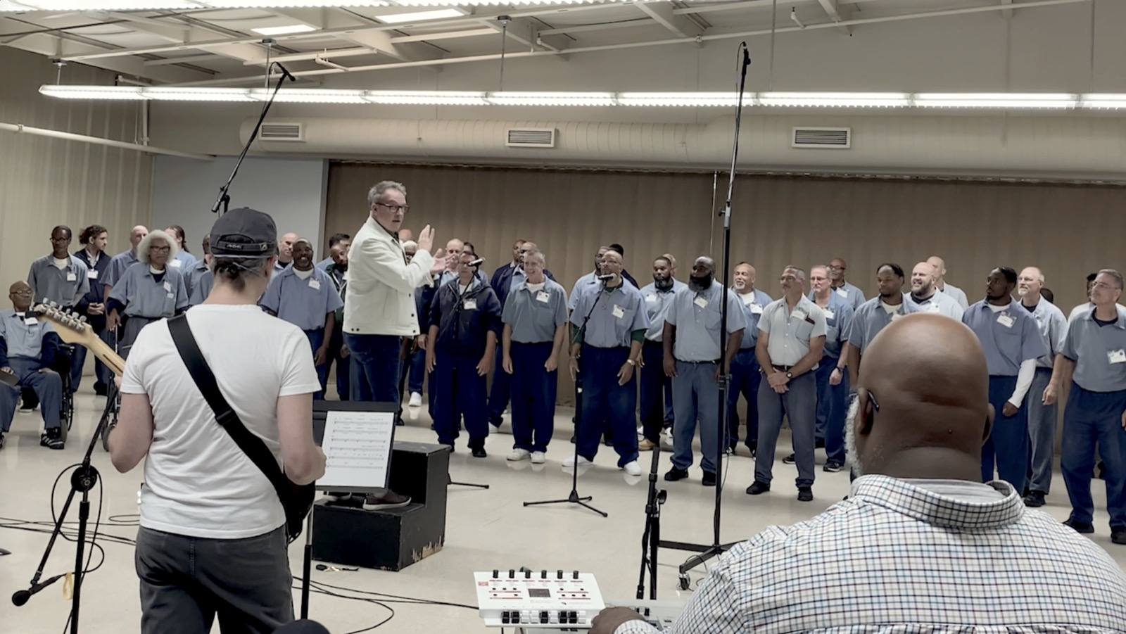 A conductor leads the standing choir while a guitarist and a drummer play in the foreground.