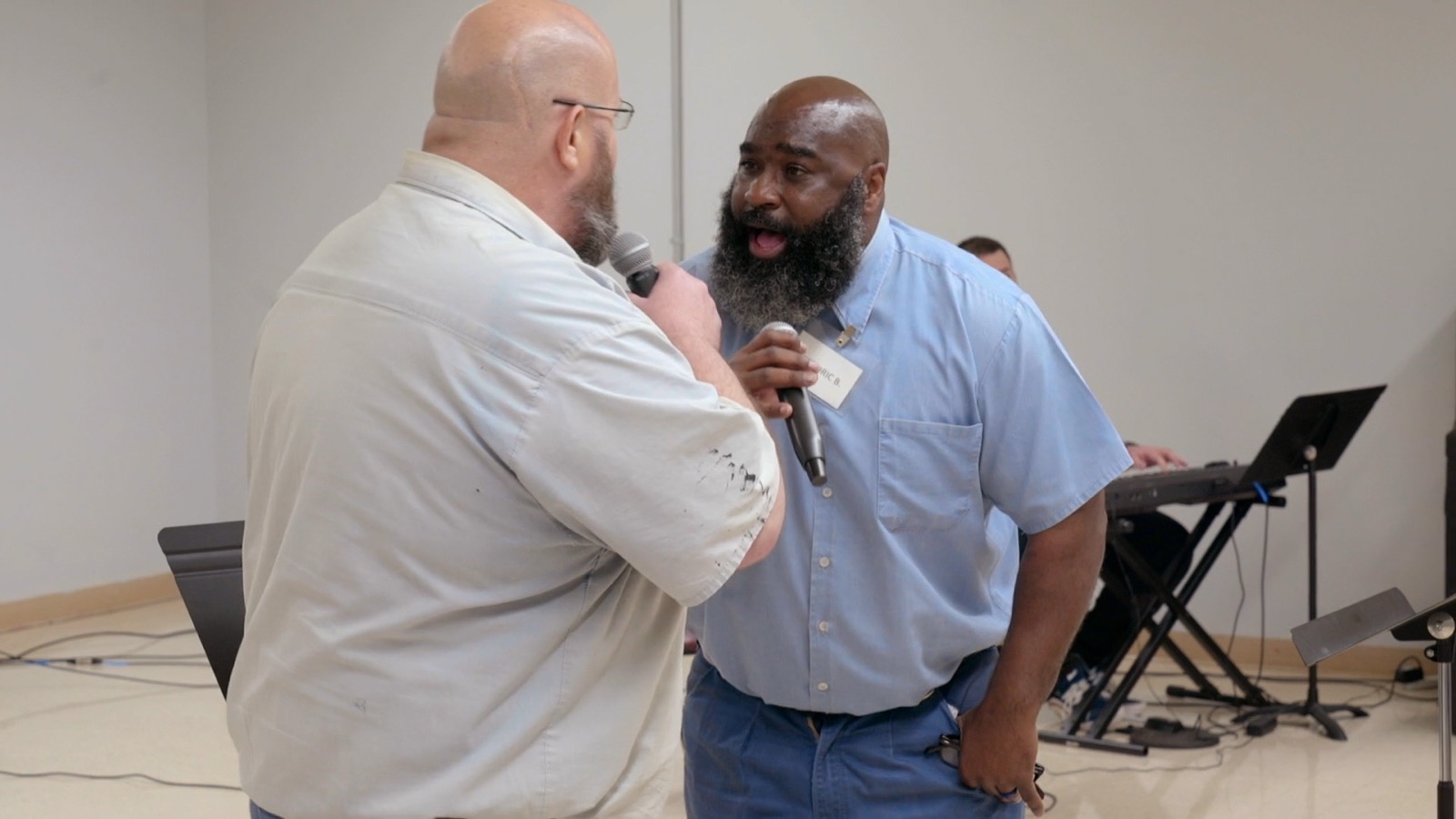 Two choir members stand face to face, one holding a microphone, singing to each other in a rehearsal room.