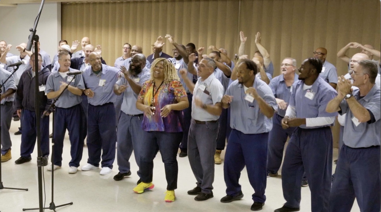 A large group singing with hands raised and clapping, a woman in a bright patterned top dancing in the middle.